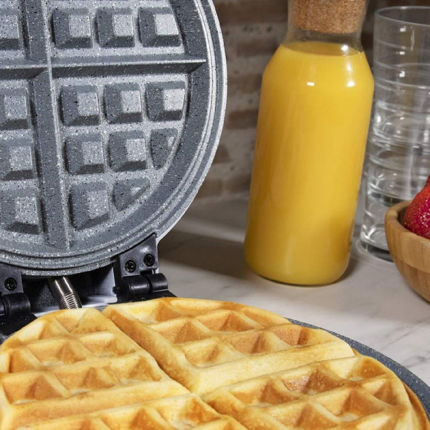 Ein Waffeleisen mit zwei goldenen Waffeln und einem Krug Orangensaft daneben.