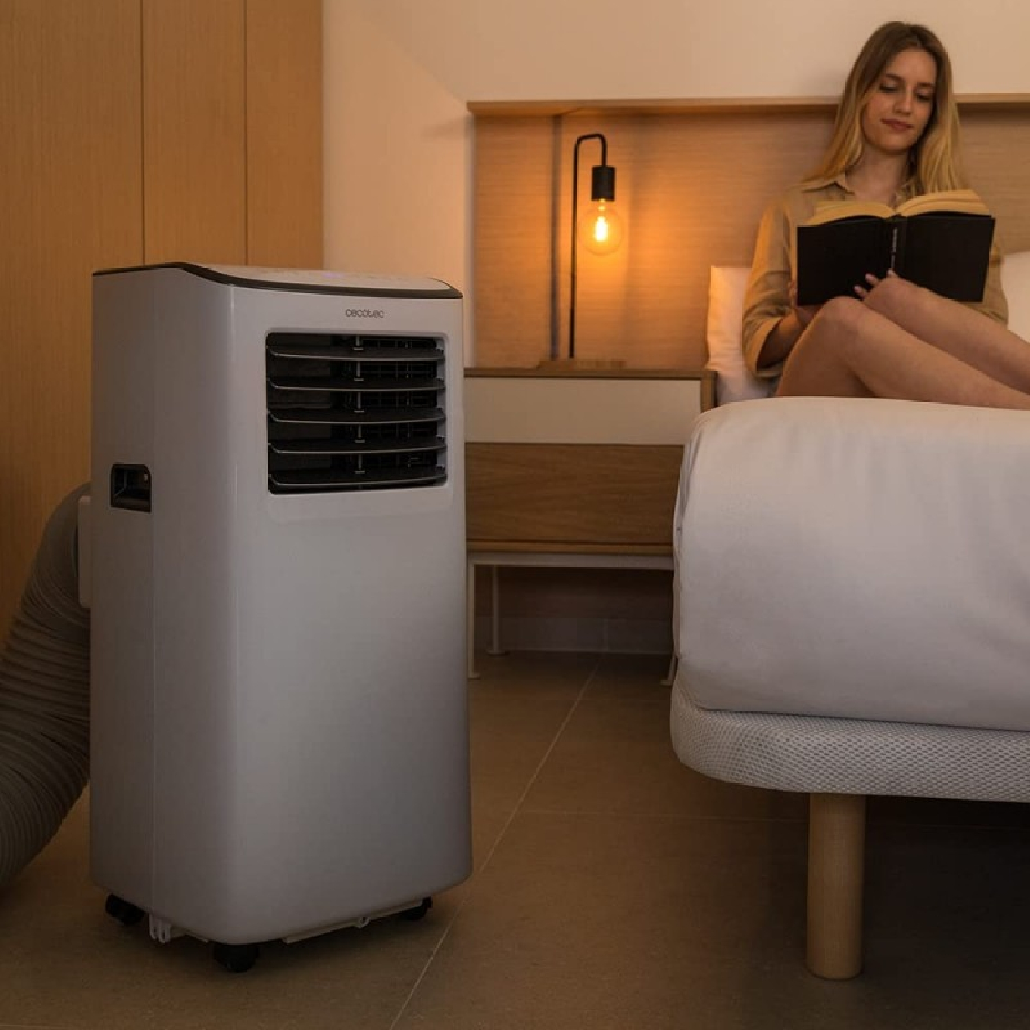 Przenośny klimatyzator stoi obok łóżka, na którym siedzi kobieta i czyta.