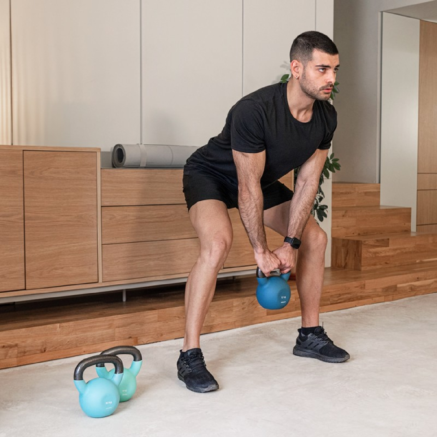 Mann in Schwarz hebt eine blaue Kettlebell. Zwei weitere Kettlebells liegen daneben.