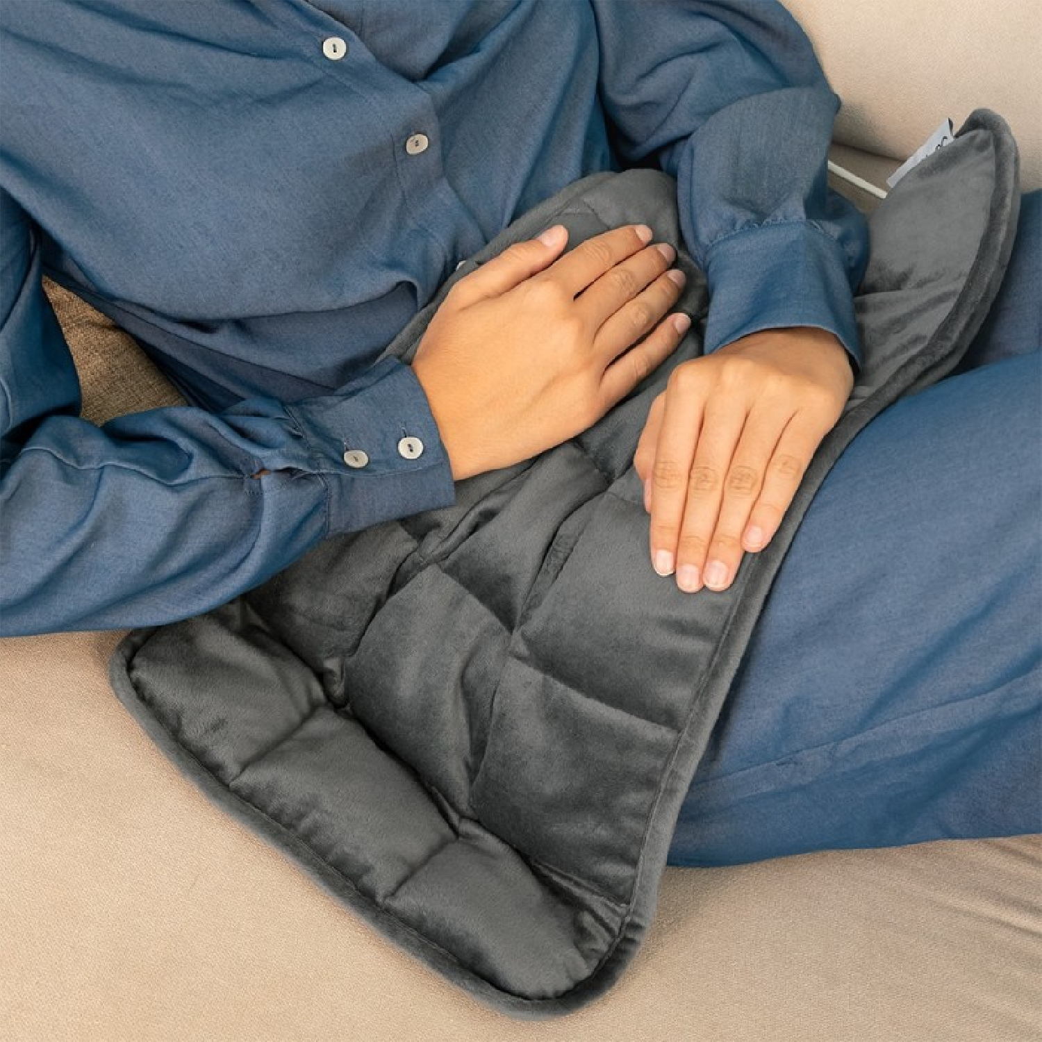 Una persona con camisa azul descansa en un sofá, sosteniendo una manta oscura acolchada.