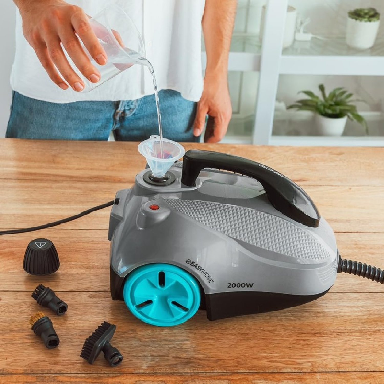 Una persona vierte agua en una limpiadora a vapor gris con accesorios sobre una mesa de madera.