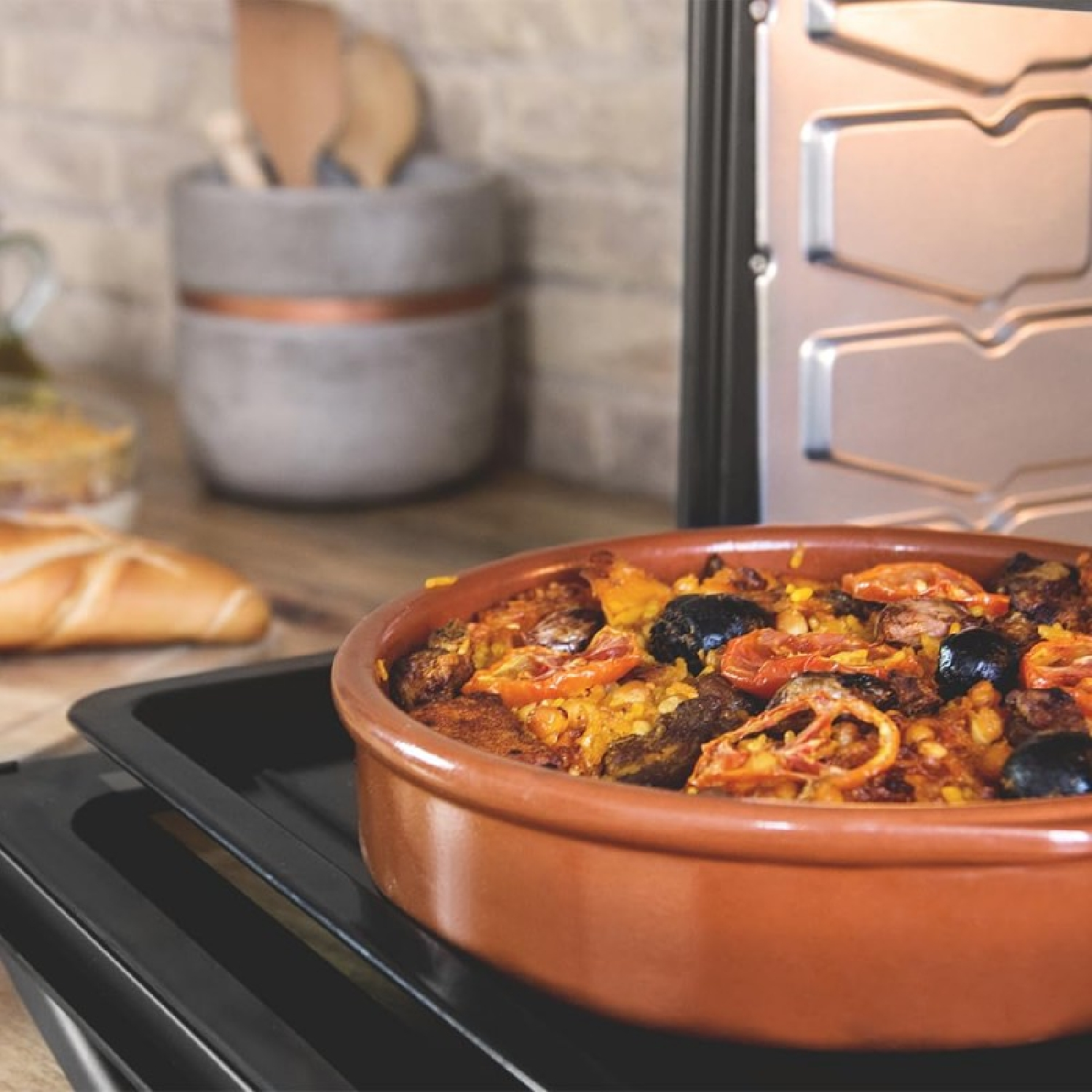 Un piatto di terracotta di paella con olive e pomodori si trova su un vassoio nero.