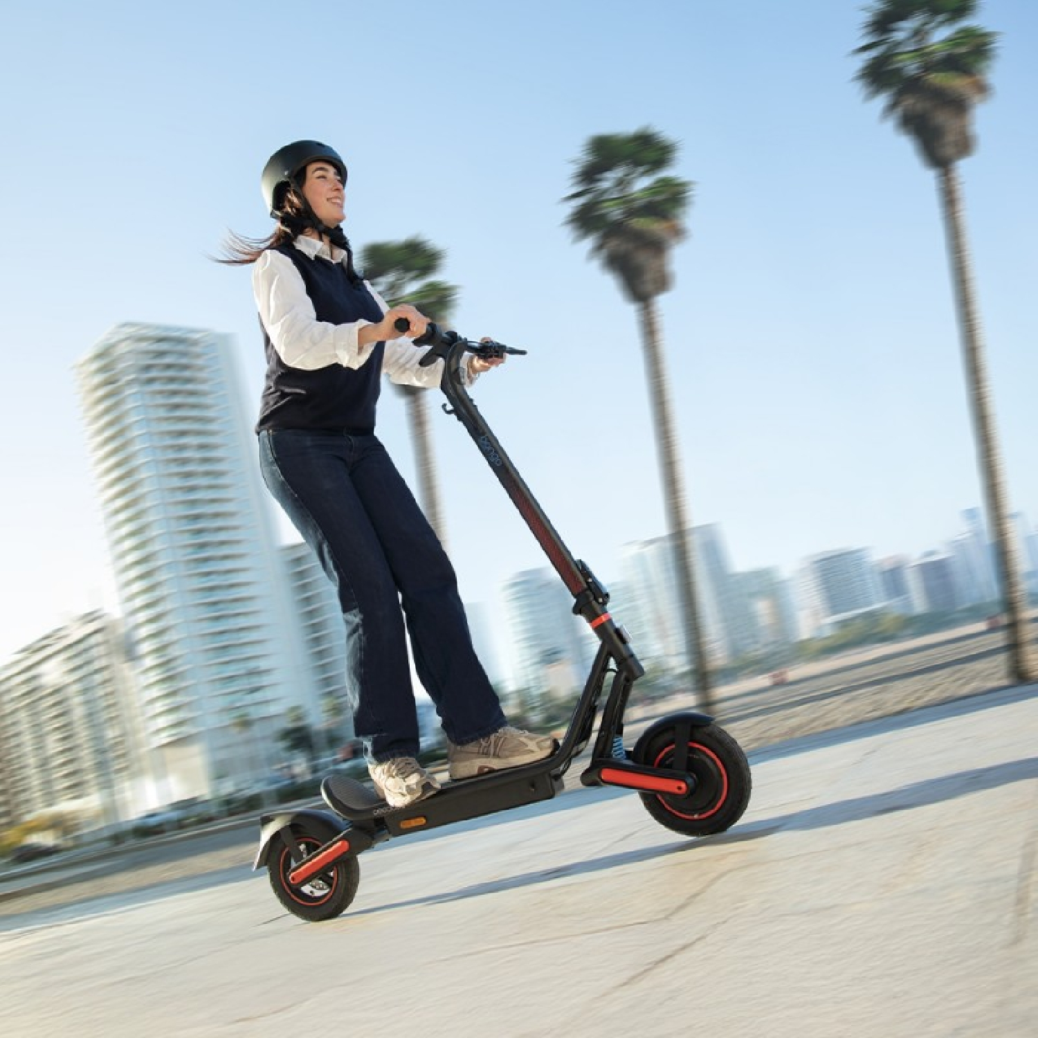 Eine Frau fährt einen E-Scooter mit Helm, Palmen und Gebäude im Hintergrund.