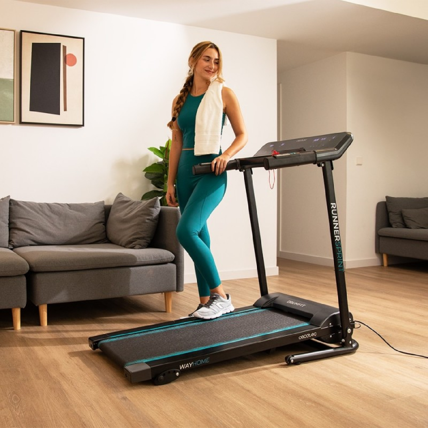Una donna in abbigliamento sportivo turchese sta accanto a un tapis roulant con un asciugamano.