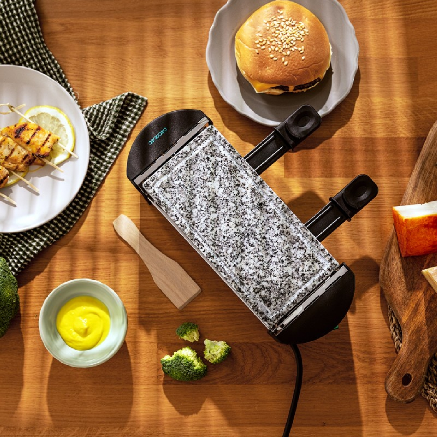 Ein Raclette-Grill mit Hähnchenspießen, Burger, Brokkoli und Dip auf einem Holztisch.