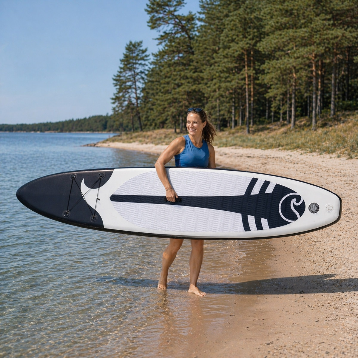Kobieta w niebieskim topie niesie biało-czarną deskę paddleboard na plaży.