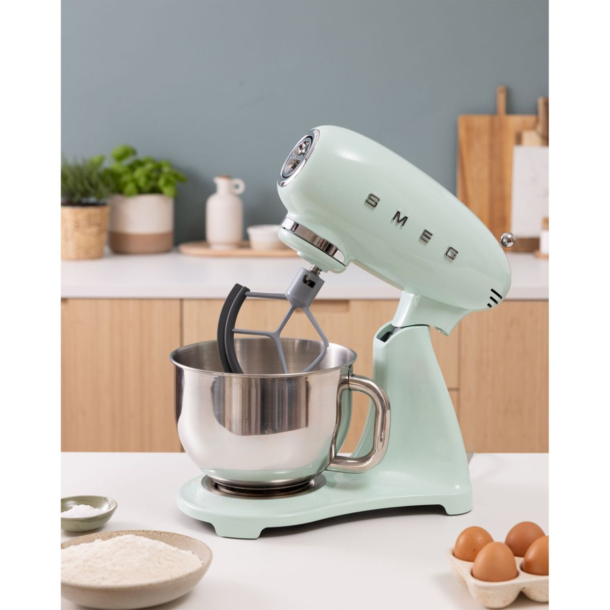 Un robot de cocina Smeg verde claro con bol, junto a harina y huevos.