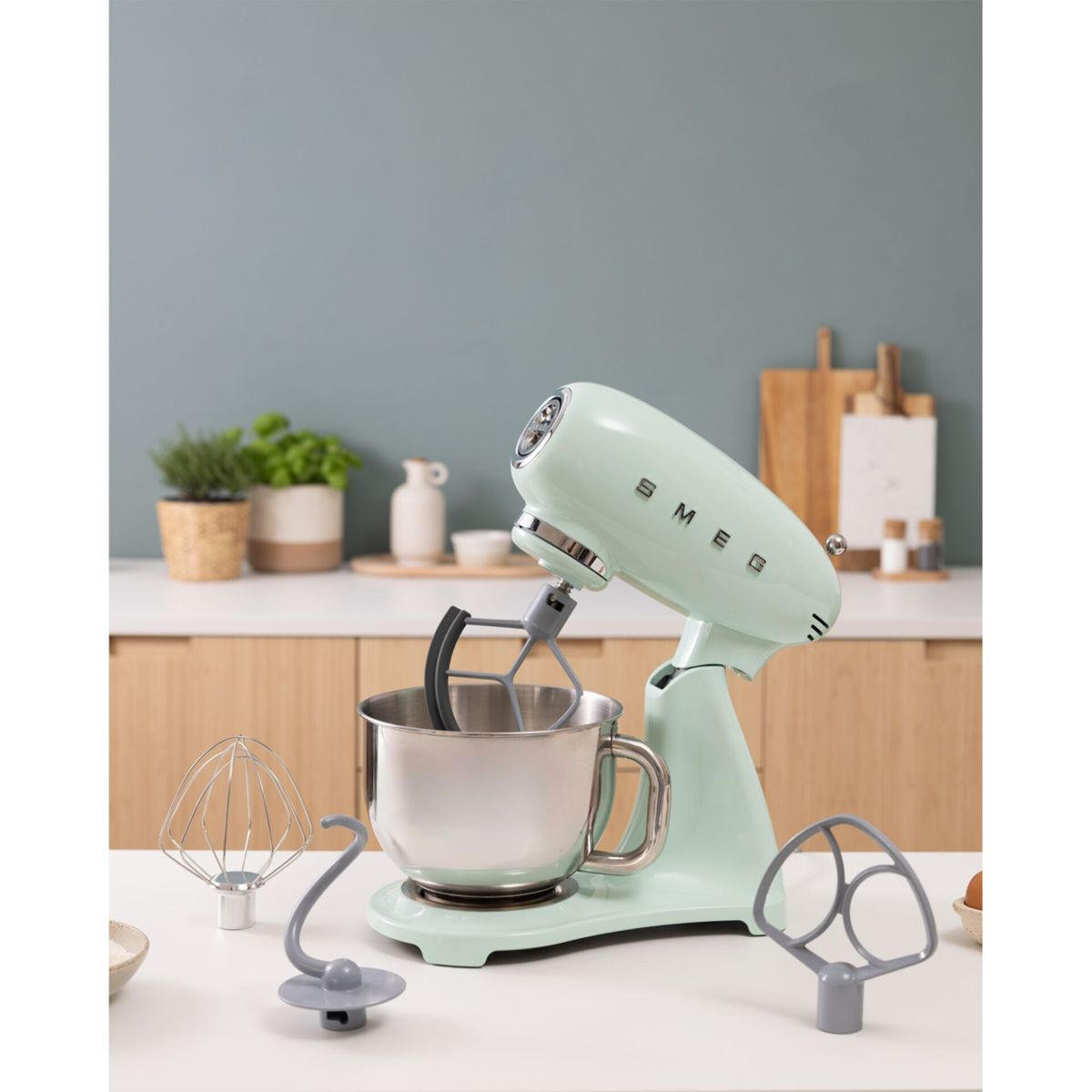 Un batidora de pie Smeg verde claro con bol y accesorios sobre una encimera.