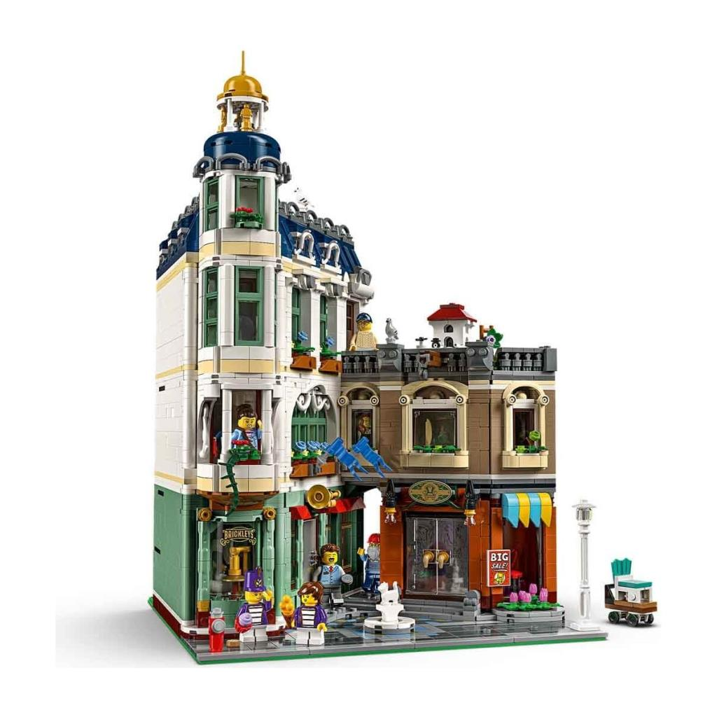 Beyaz arka plan üzerinde köşe kulesi, dükkanlar ve minfigürler içeren detaylı bir Lego binası.
