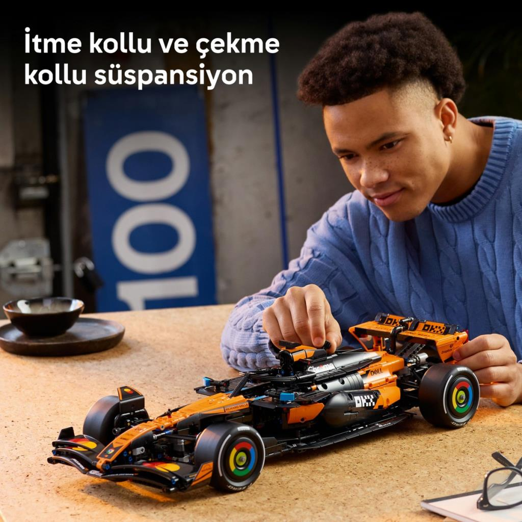Genç bir adam ahşap bir masada detaylı, turuncu bir Lego yarış arabası yapıyor.