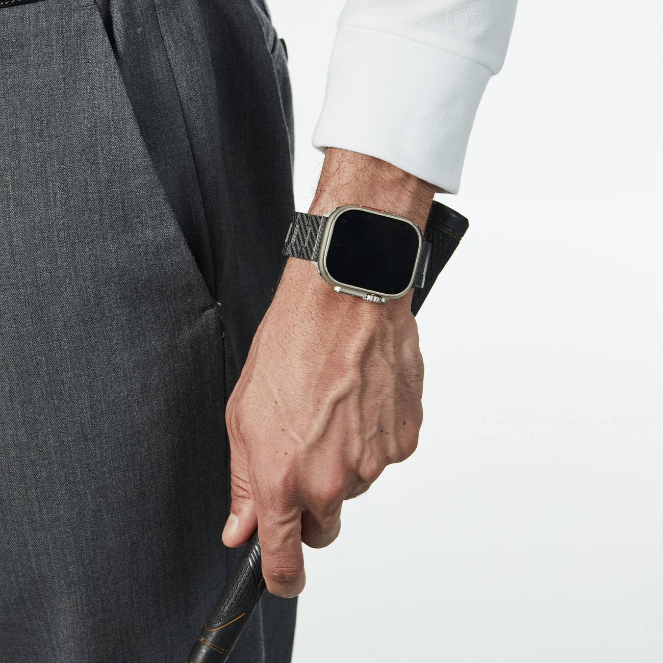 Un hombre con camisa blanca y pantalones grises sostiene un palo de golf, lleva un smartwatch.