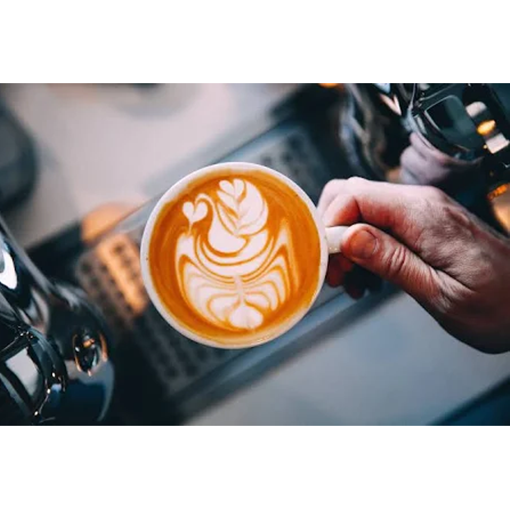 Eine weiße Tasse mit Latte Art, gehalten von einer Hand, mit Kaffeemaschinenteilen im Hintergrund.