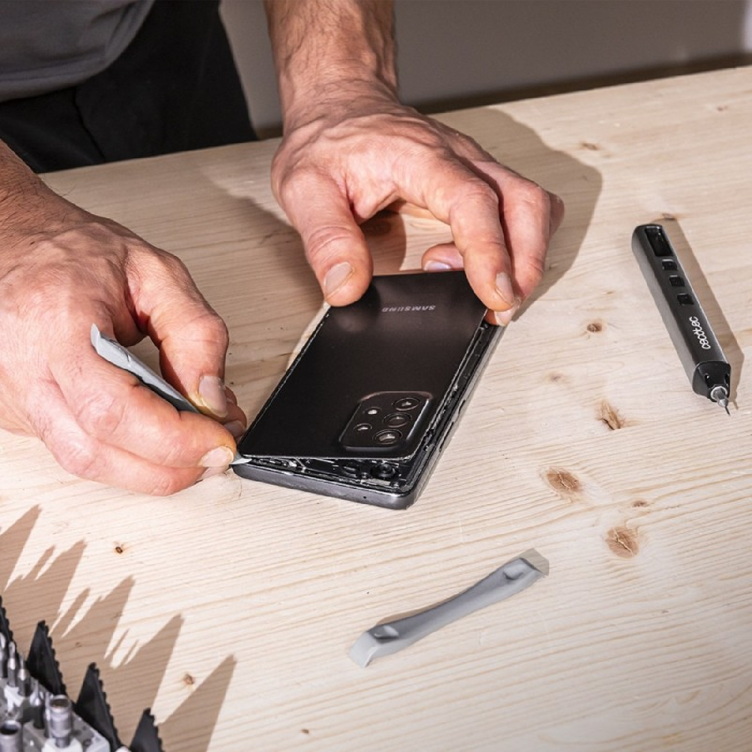 Manos usan herramientas para abrir un teléfono Samsung negro en una mesa de madera.