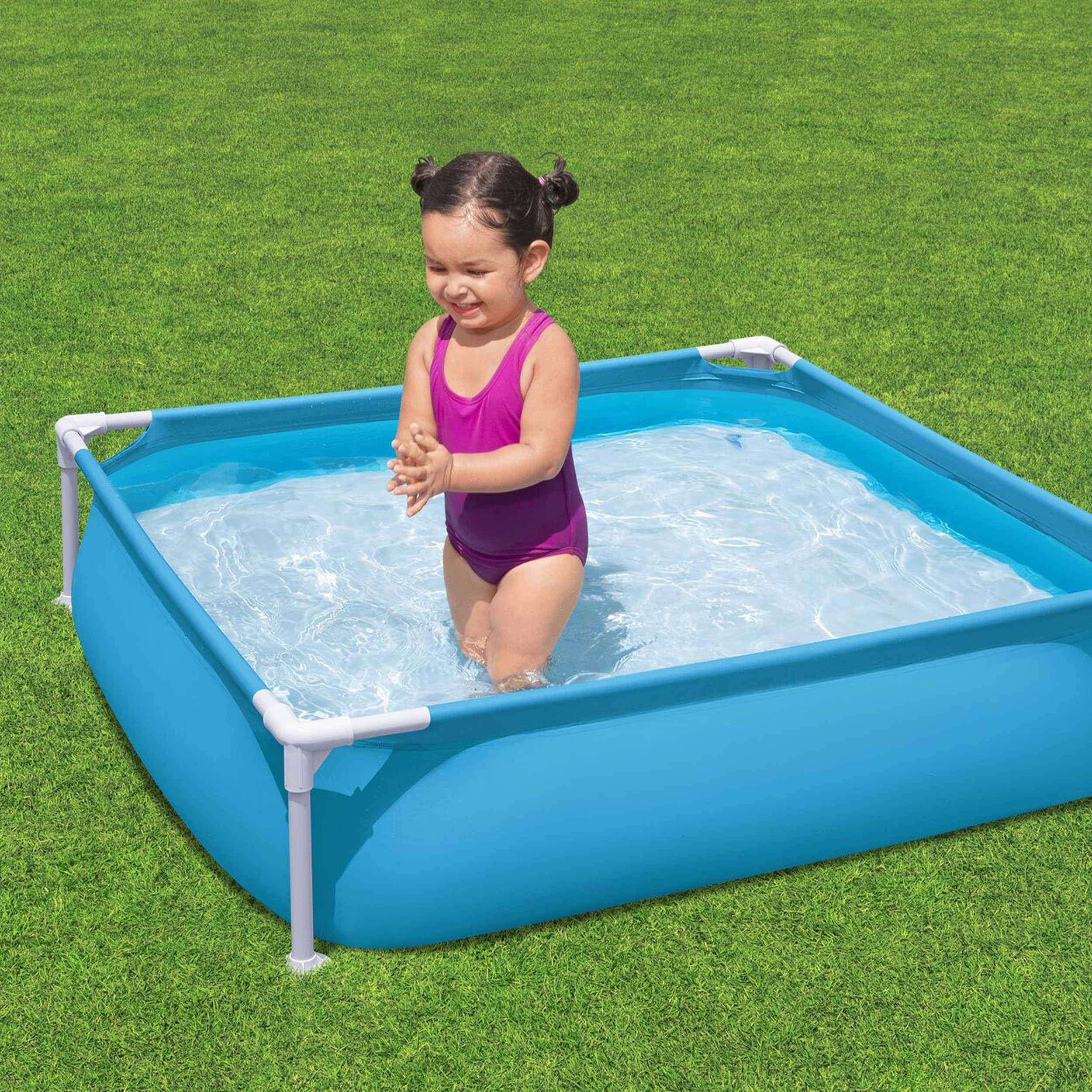 Mała dziewczynka w fioletowym stroju kąpielowym stoi w niebieskim brodziku na zielonej trawie.