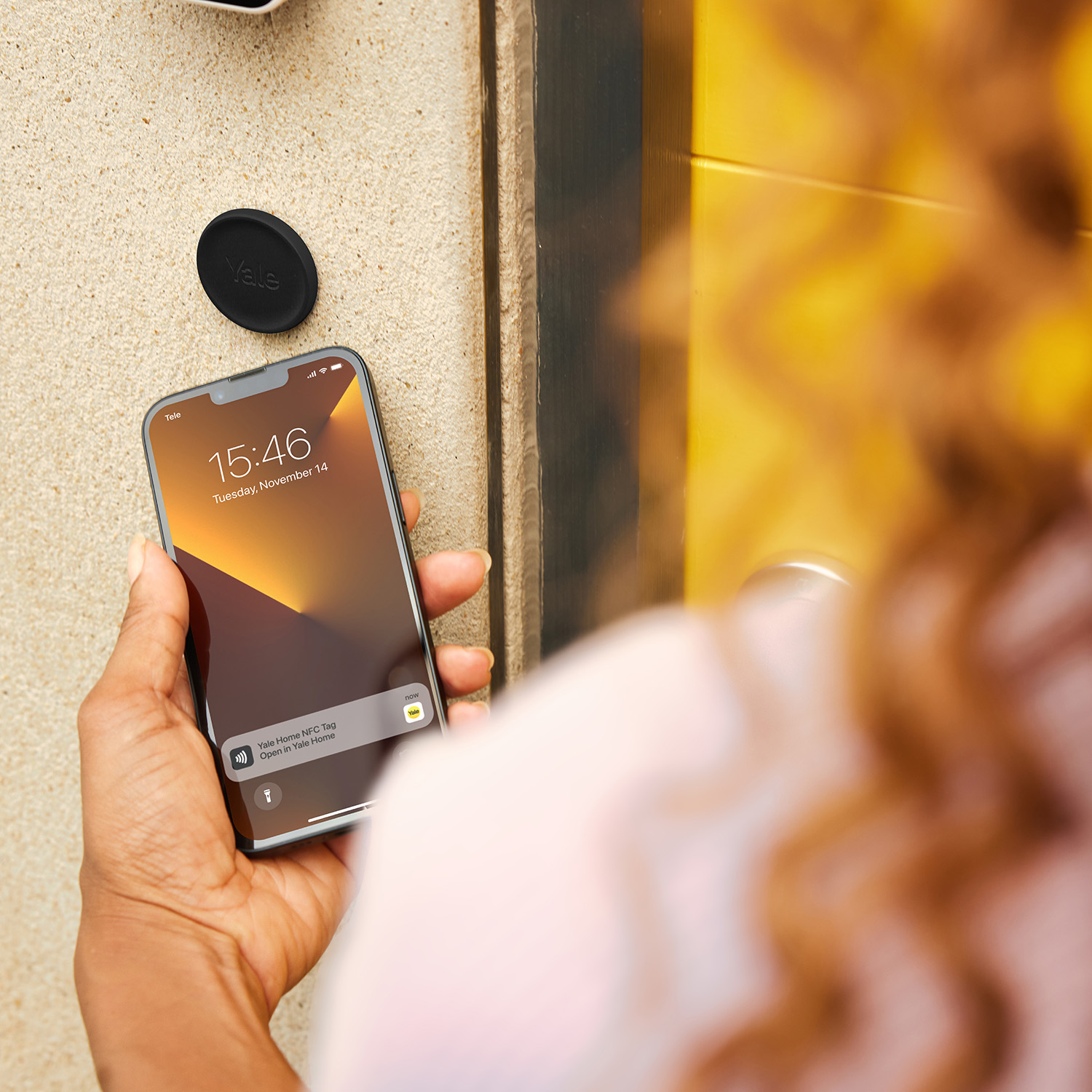 Una mano sujeta un smartphone cerca de una pared con una etiqueta Yale. El teléfono muestra una notificación.