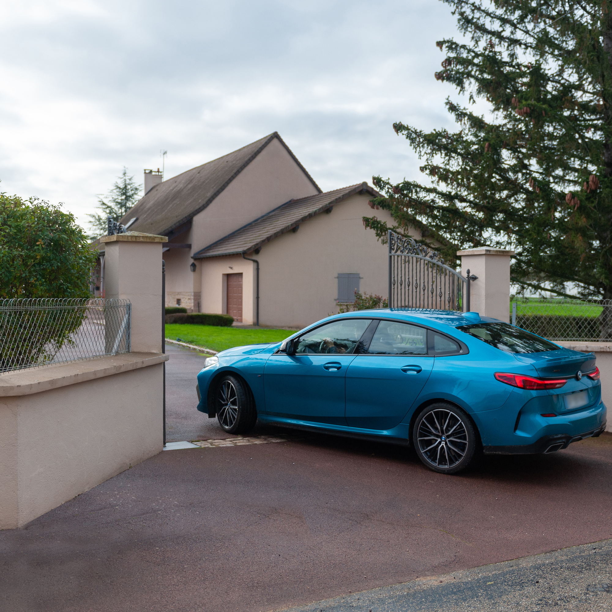 Un BMW azul conduce por un camino de entrada hacia una casa, rodeado de árboles y una puerta.