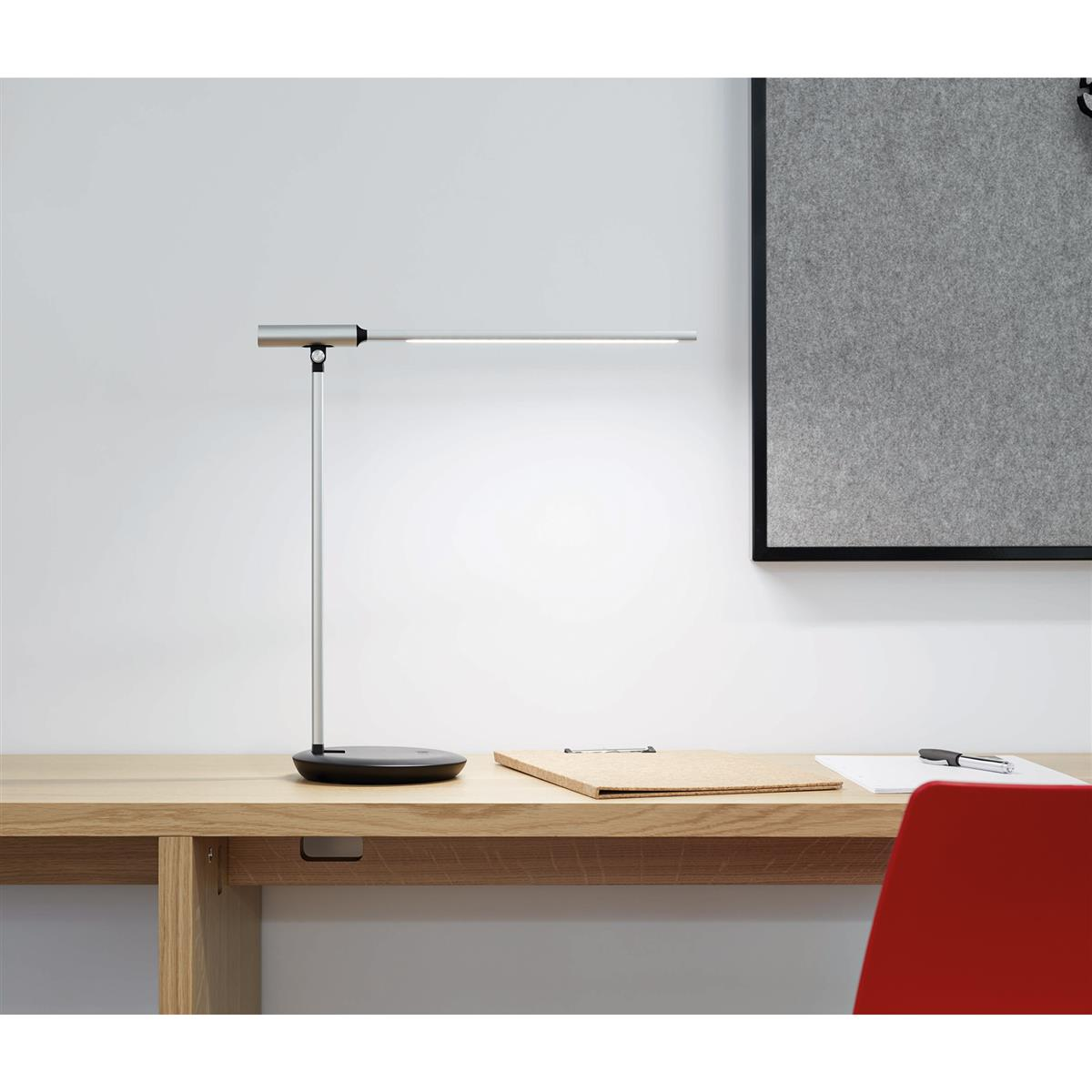 Un brazo de escritorio plateado con luz LED se asienta sobre un escritorio de madera junto a un tablón de anuncios.