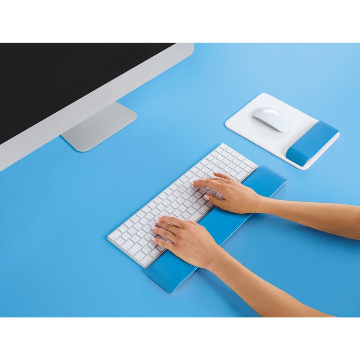Manos escribiendo en un teclado blanco con reposamuñecas azules sobre un escritorio azul.