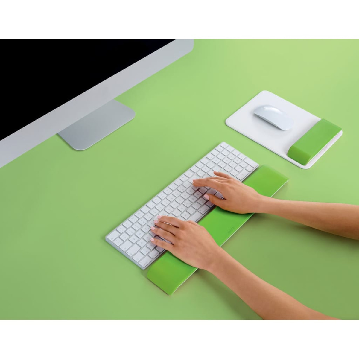 Manos escribiendo en un teclado con reposamuñecas verdes, junto a un monitor.
