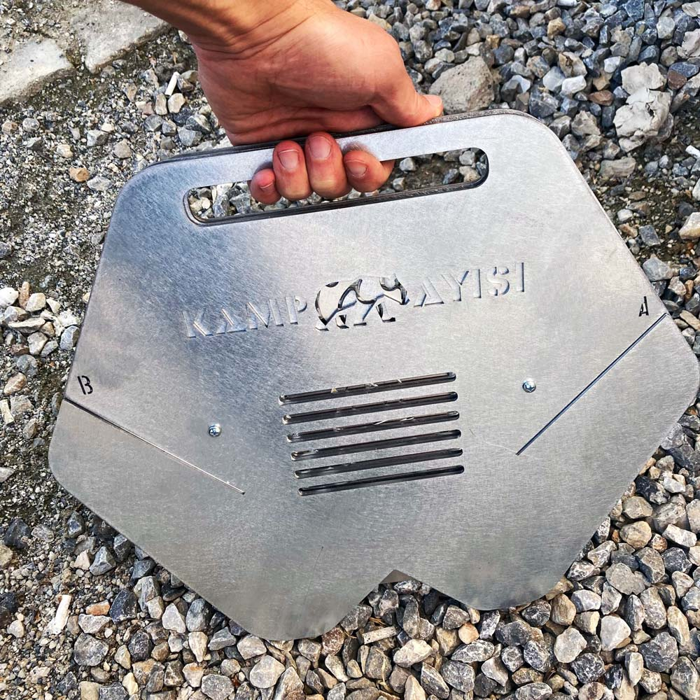 El, 'KAMPAYISI' logolu ve ızgara havalandırmalı metal bir nesneyi çakıl üzerinde tutuyor.