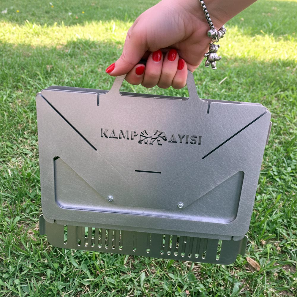 Kırmızı ojeli bir el, yeşil çimenlerin üzerinde katlanabilir metal mangal tutuyor.