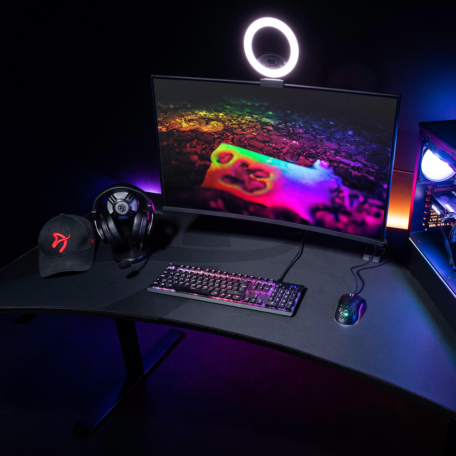 Een gaming bureau-opstelling met een gebogen monitor, toetsenbord, muis, koptelefoon, pet en pc-kast.
