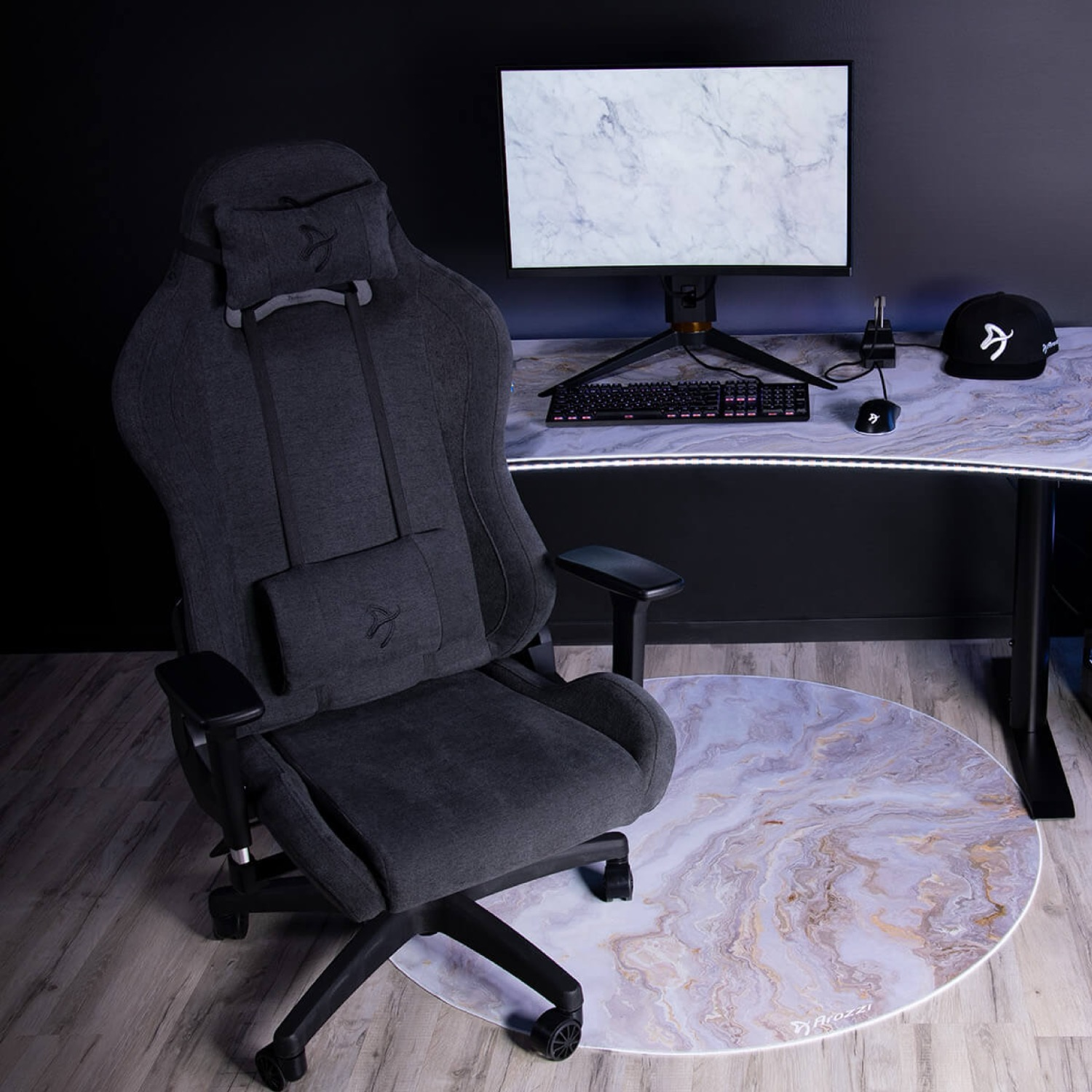 Ein dunkelgrauer Gaming-Stuhl steht auf einer Marmormuster-Bodenmatte. Ein Schreibtisch mit Monitor, Tastatur, Maus & Kappe.
