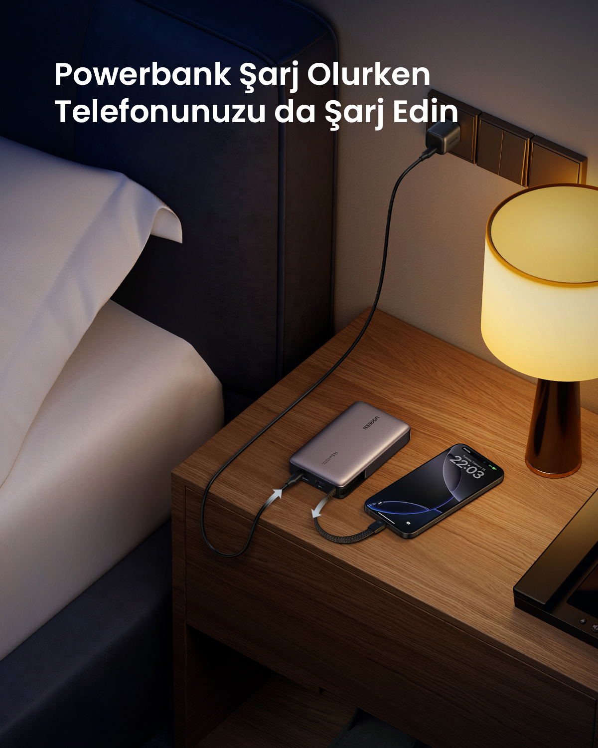 Bir powerbank, komodinin üzerindeki bir telefonu şarj ediyor, yanında lamba var.