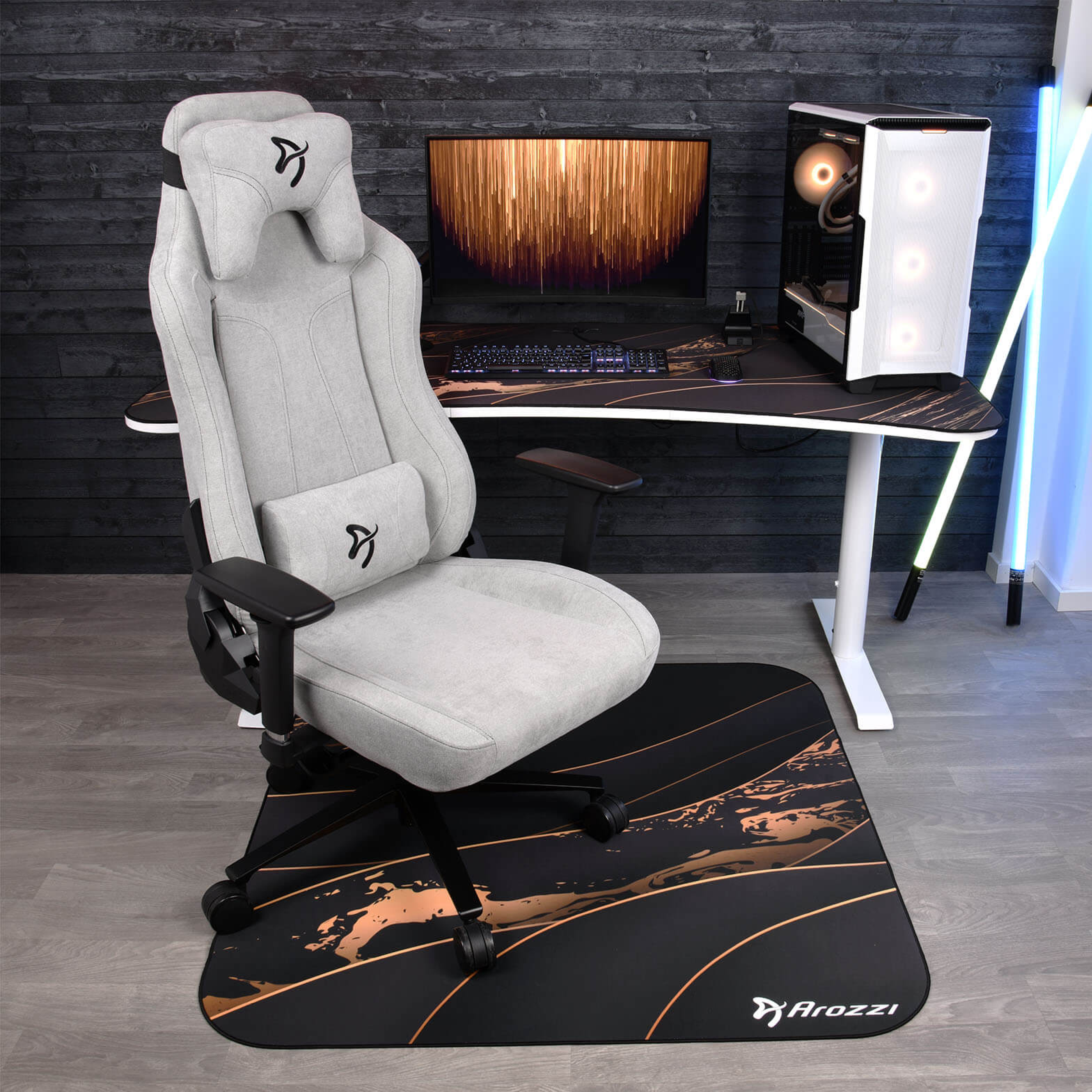 Ein grauer Gaming-Stuhl steht auf einer schwarzen Matte mit Kupferakzenten. Ein Schreibtisch mit PC, Monitor und Tastatur ist im Hintergrund zu sehen.
