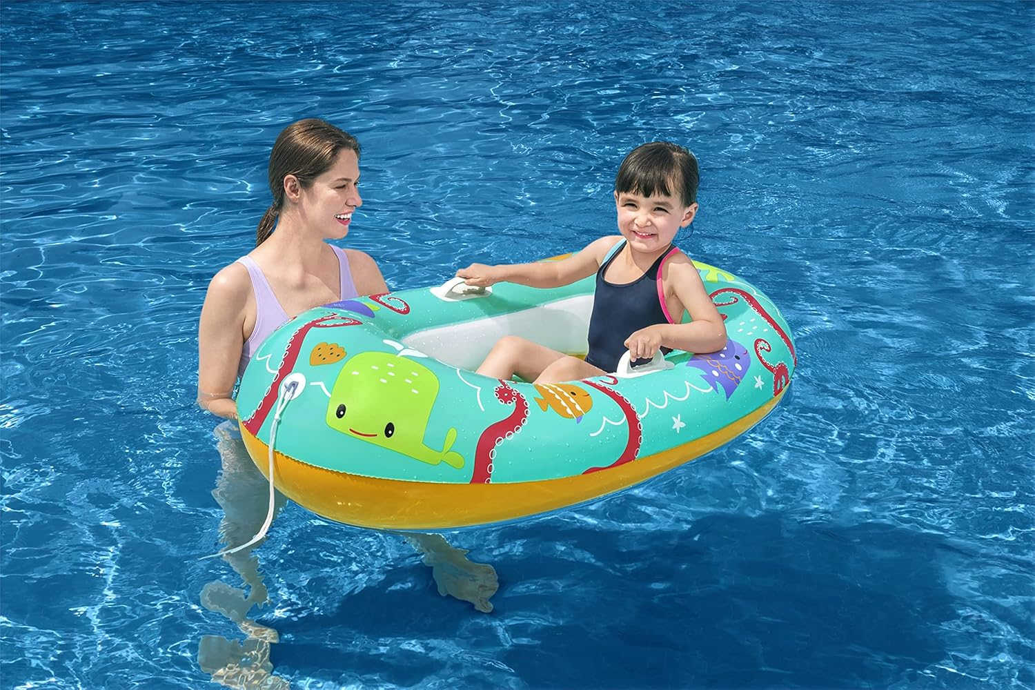 Ein Mädchen sitzt in einem aufblasbaren Boot mit Wal- und Oktopus-Motiv, während eine Frau daneben im Pool steht.