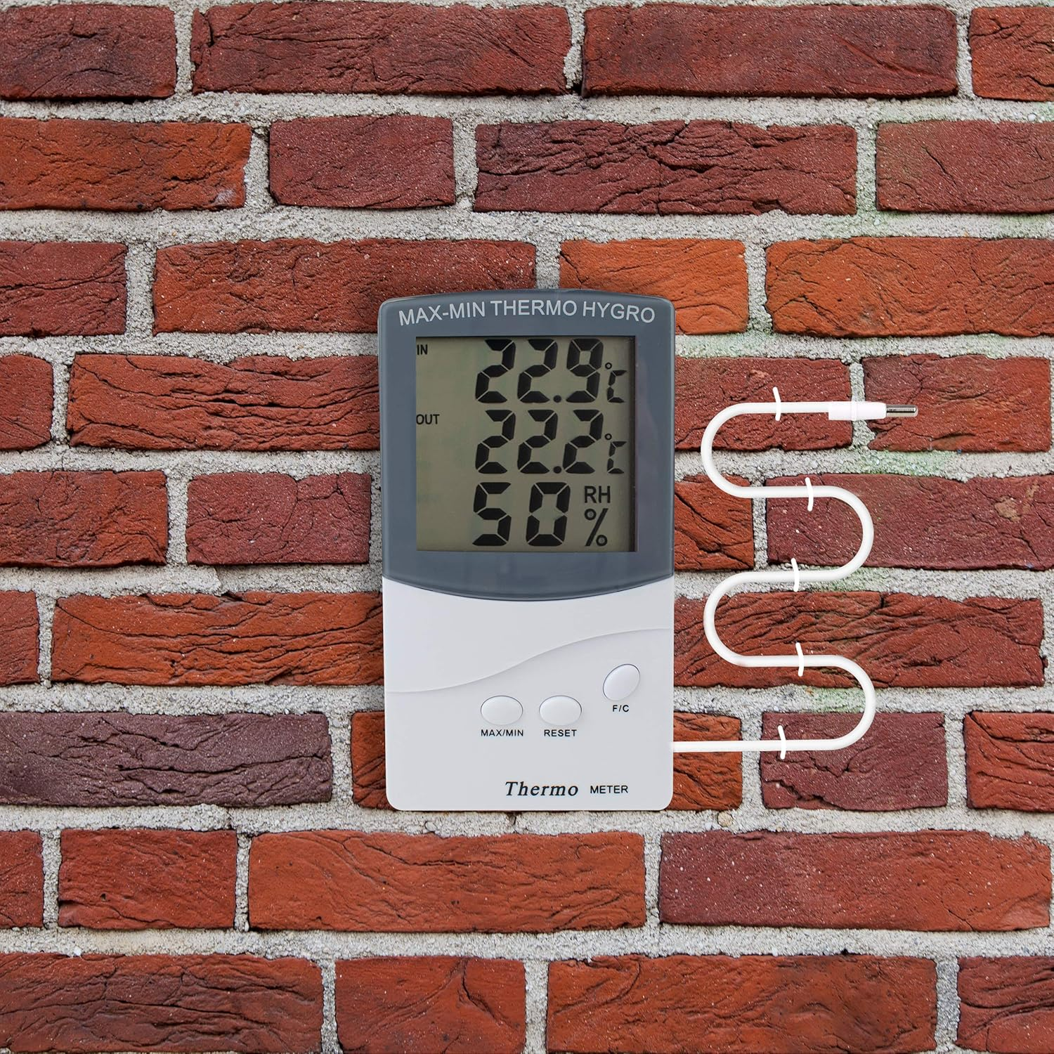Ein digitales Thermo-Hygrometer mit Kabel hängt vor einer Ziegelwand.