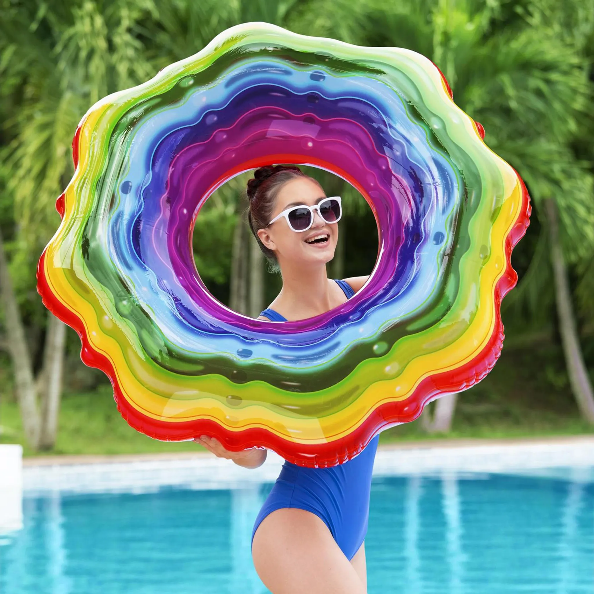 Ein Mädchen mit Sonnenbrille hält einen bunten Schwimmring vor einem Pool.