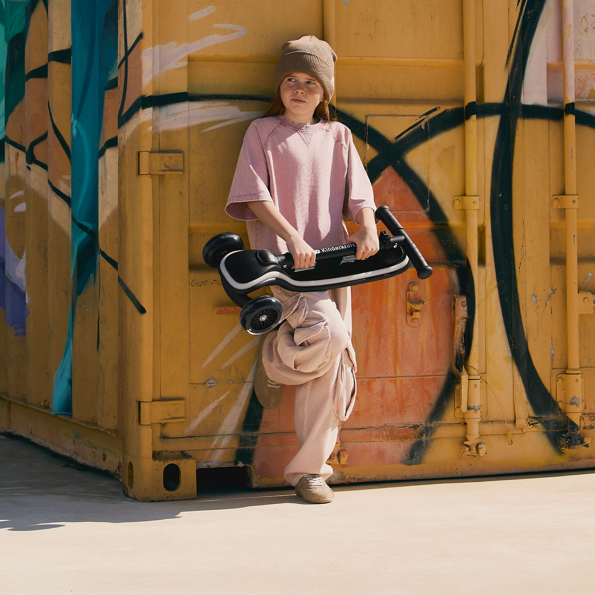 Ein Mädchen mit Beanie und rosafarbenem Shirt hält einen Roller vor einer gelben Wand mit Graffiti.