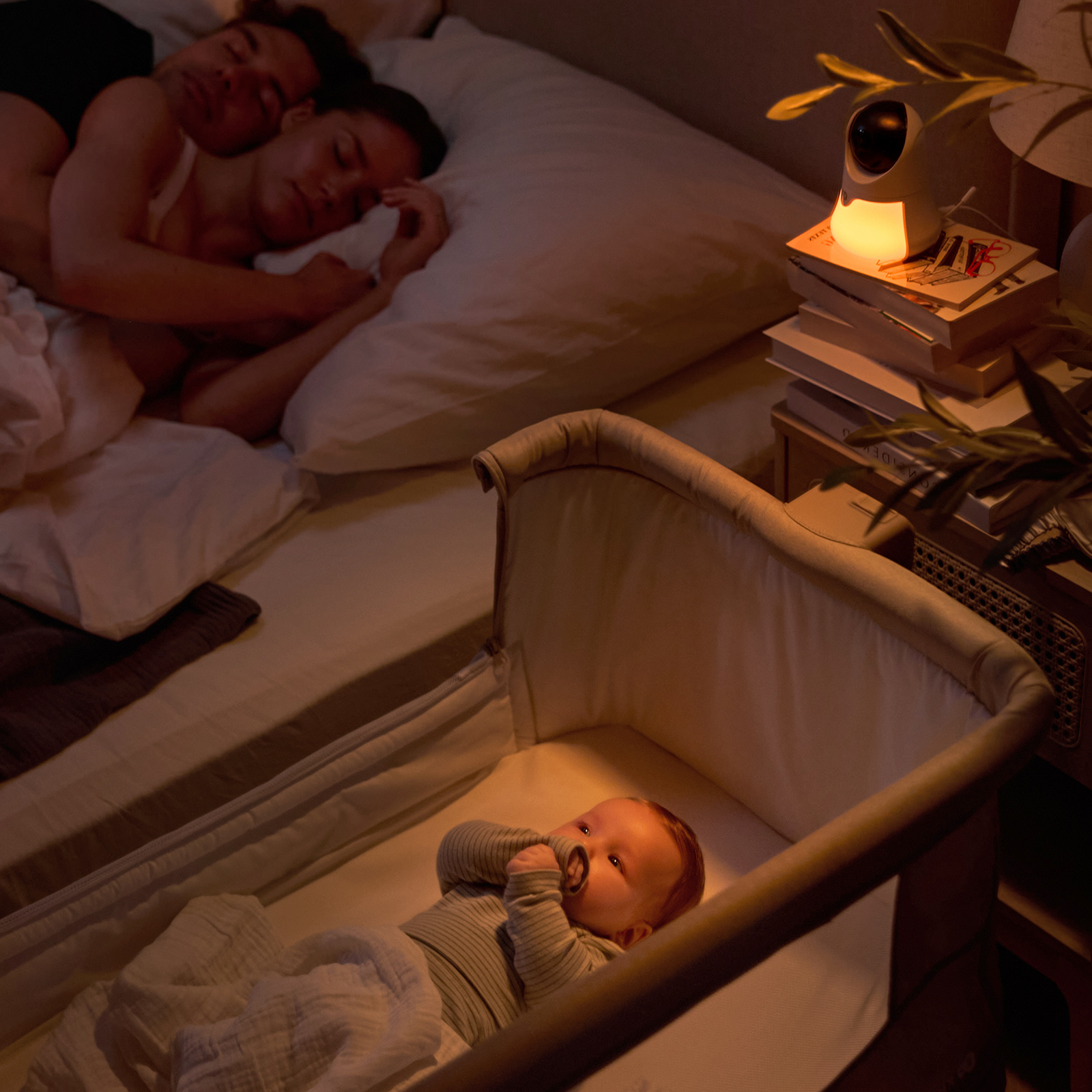 Ein Baby liegt in einem Stubenwagen, Eltern schlafen im Bett. Ein Babyphone leuchtet.