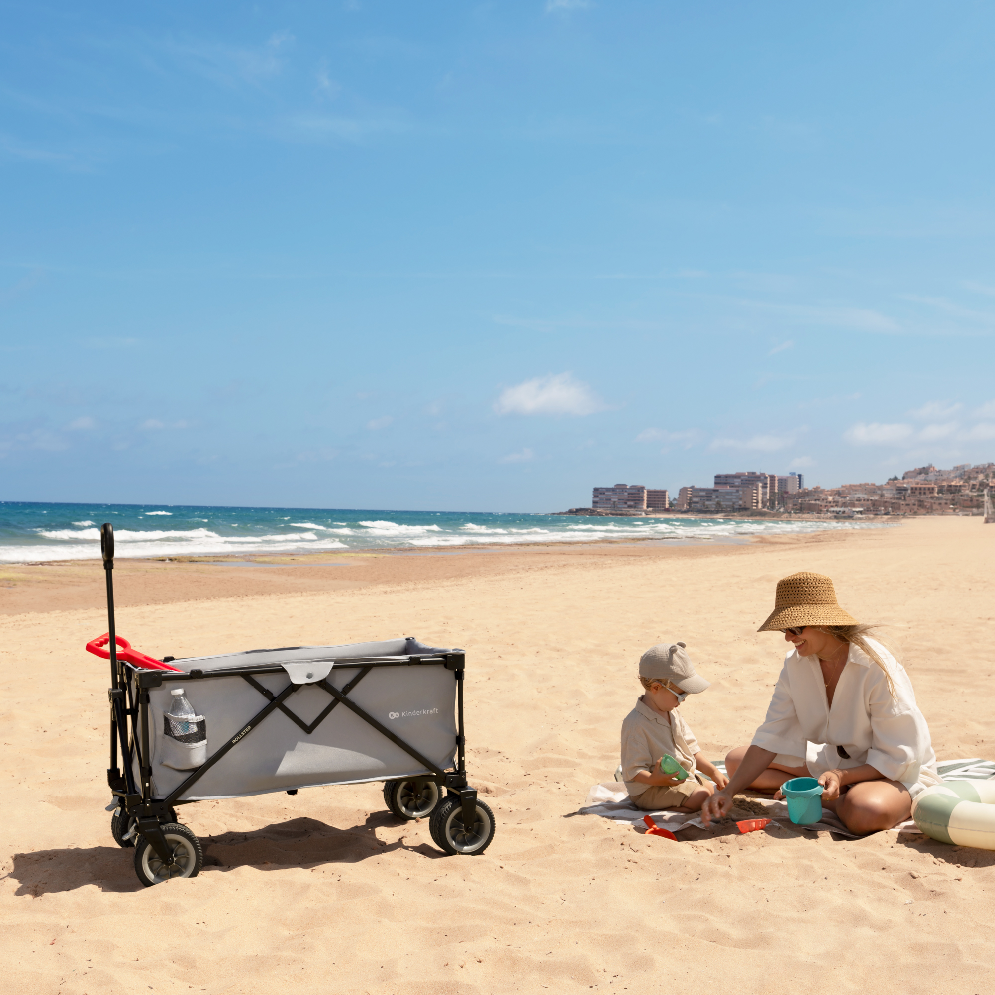 Frau und Kind spielen am Strand mit einem Bollerwagen.