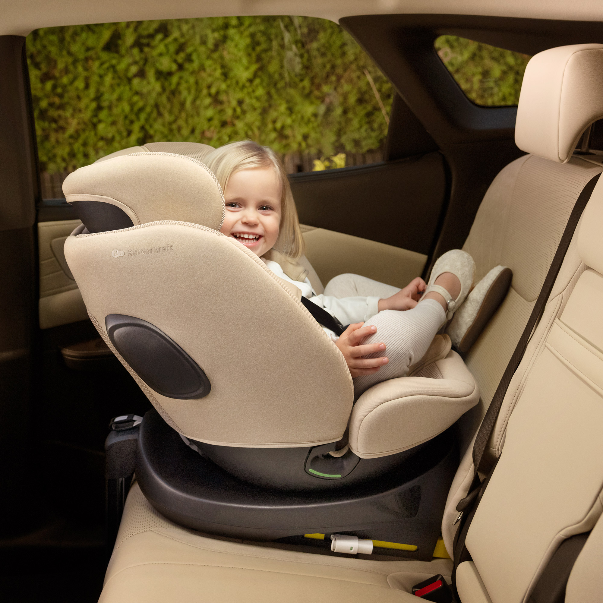 Ein fröhliches kleines Mädchen sitzt in einem beigefarbenen Kindersitz im Auto.