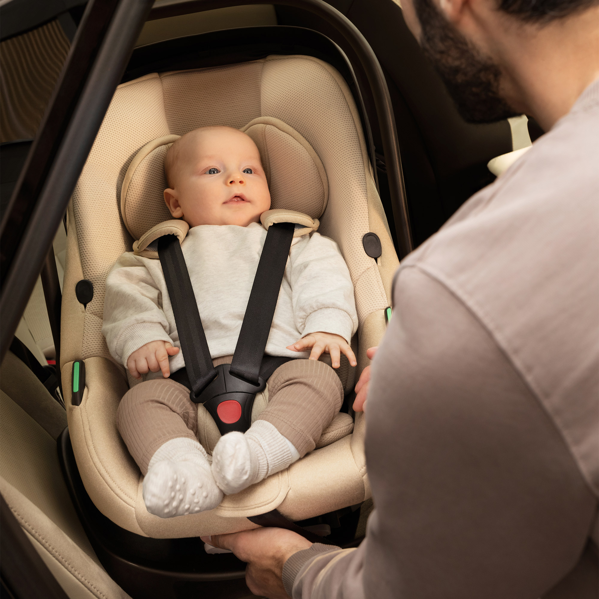 Ein Baby sitzt in einem hellbeigen Kindersitz im Auto, ein Mann hilft.
