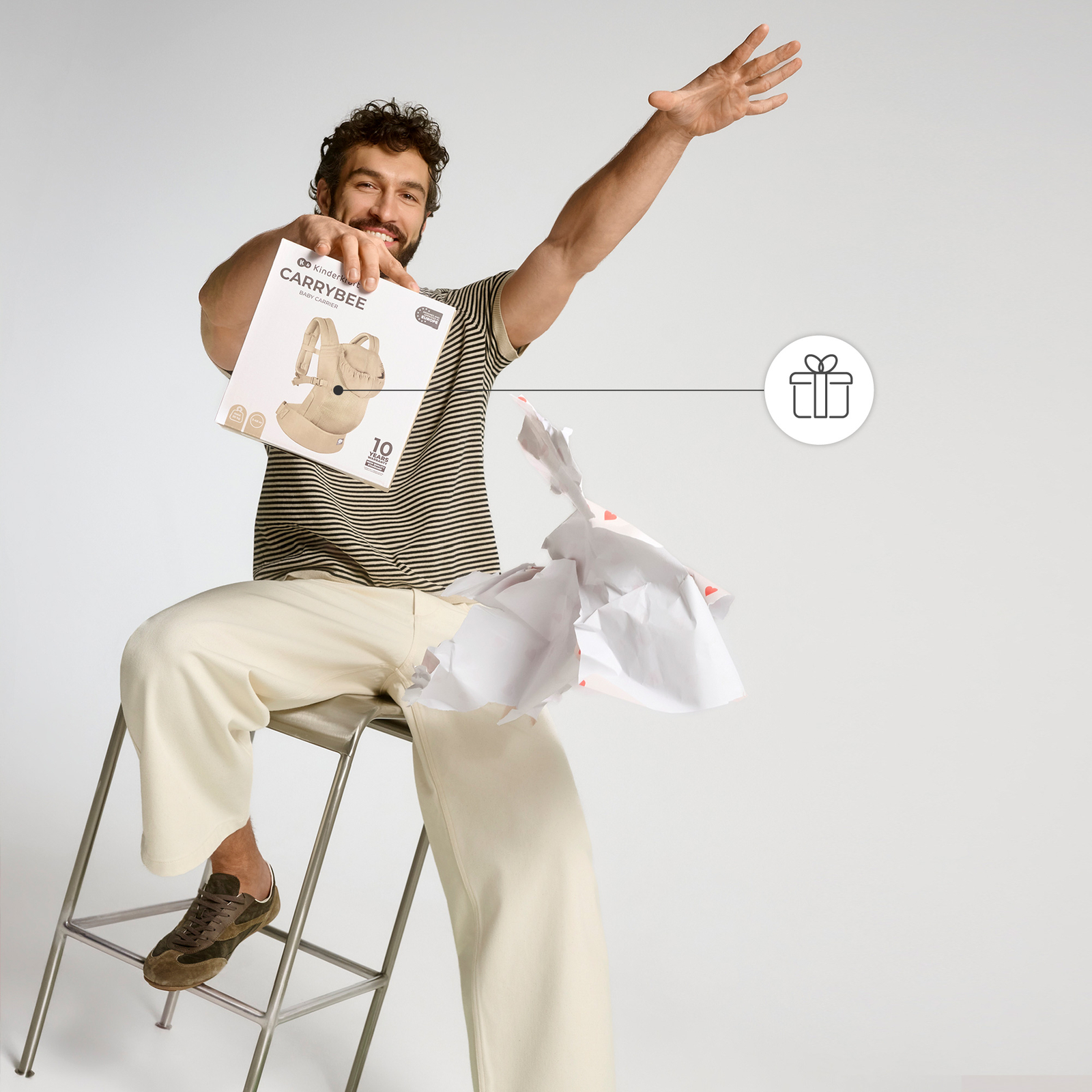 Mann sitzt auf Hocker, hält Kinderwagen-Box. Zerknülltes Papier liegt daneben.