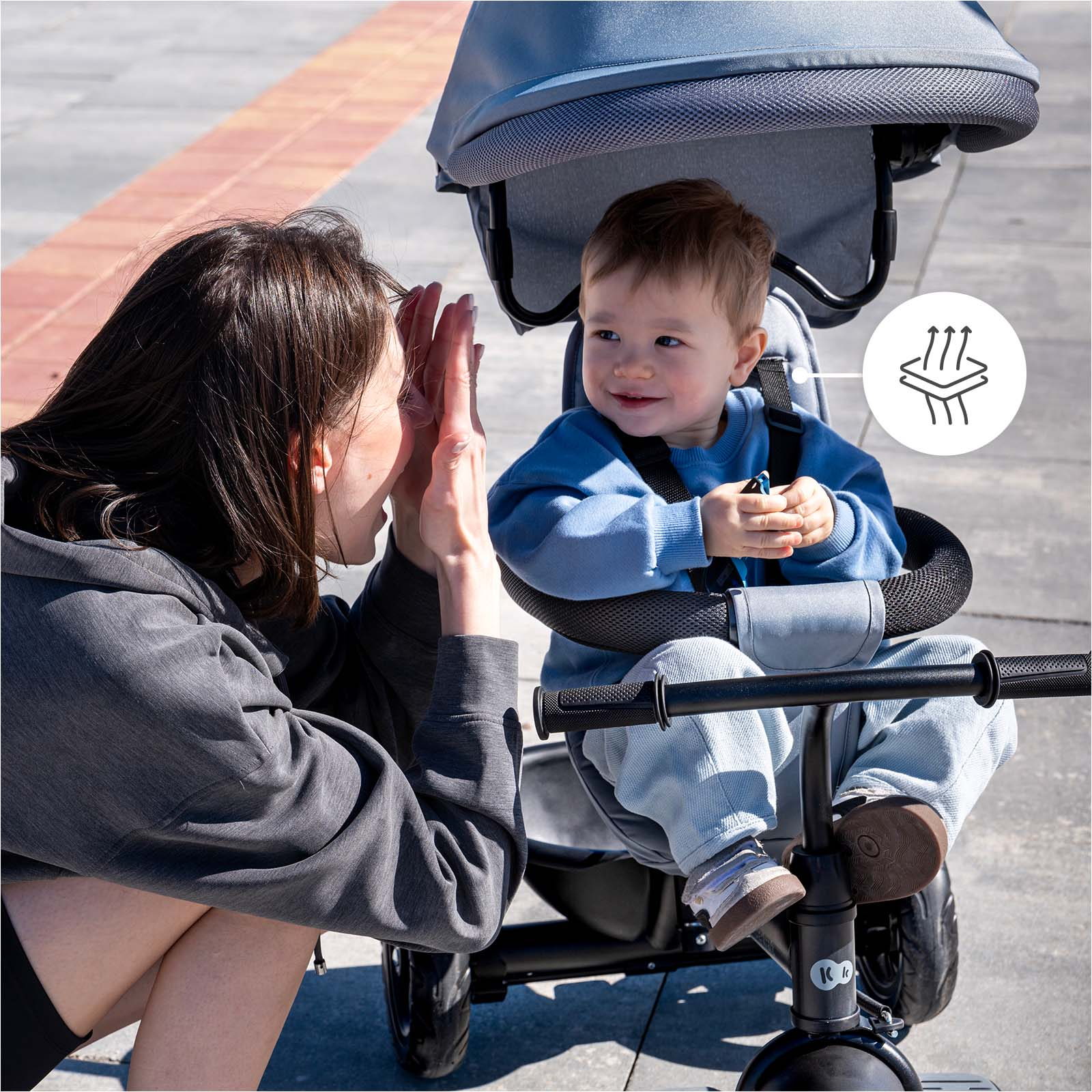Eine Frau gibt einem Baby in einem Kinderwagen High-Five. Ein Symbol für Luftzirkulation ist sichtbar.