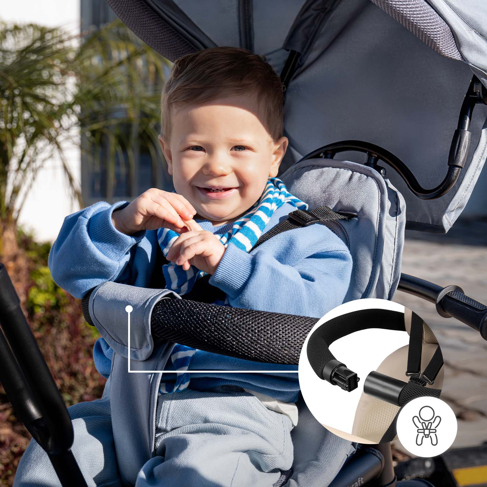 Ein lächelnder Säugling im Kinderwagen, blaue Sweatshirt, gestreifter Schal. Detailansicht des Gurtzeugs.