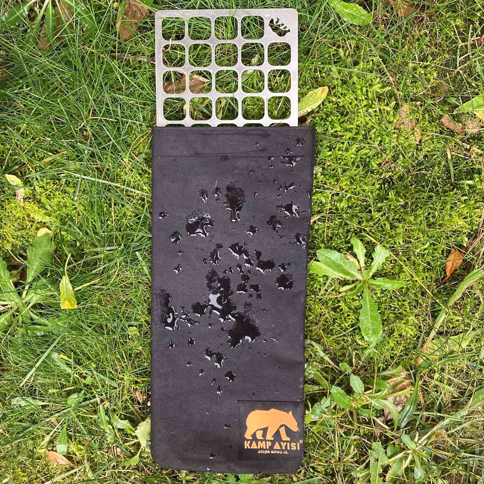 Metal plakalı siyah kumaş çanta çimenlerin üzerinde duruyor. Kumaşta su lekeleri var.