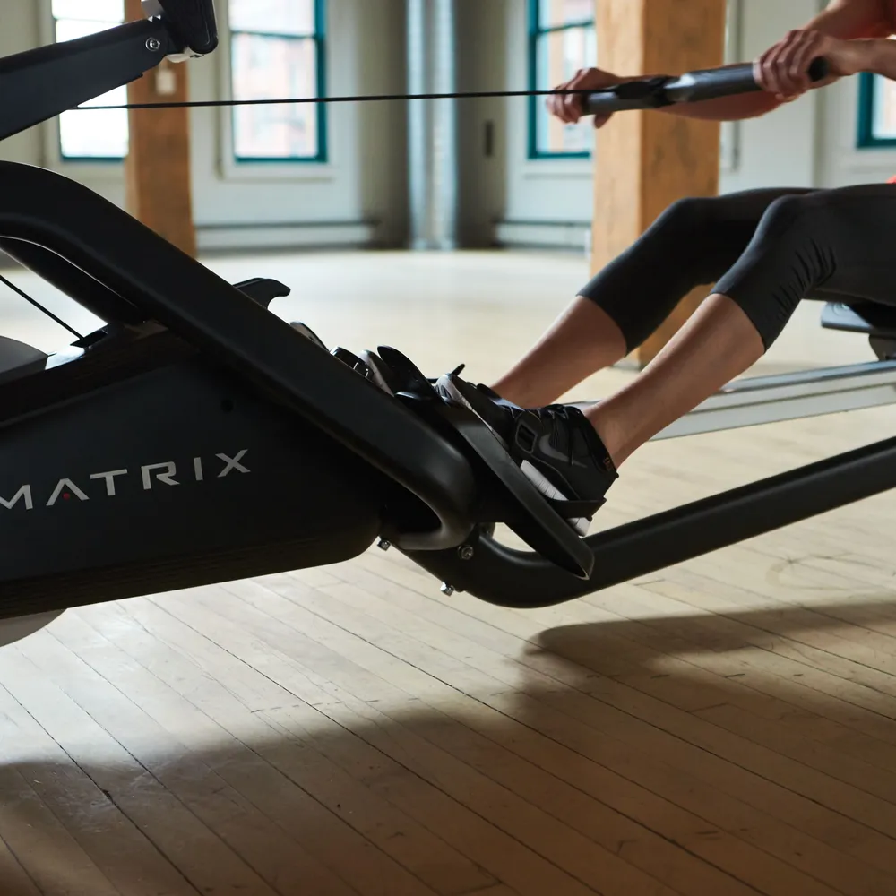 A person works out on a rowing machine in a bright room with a wooden floor.