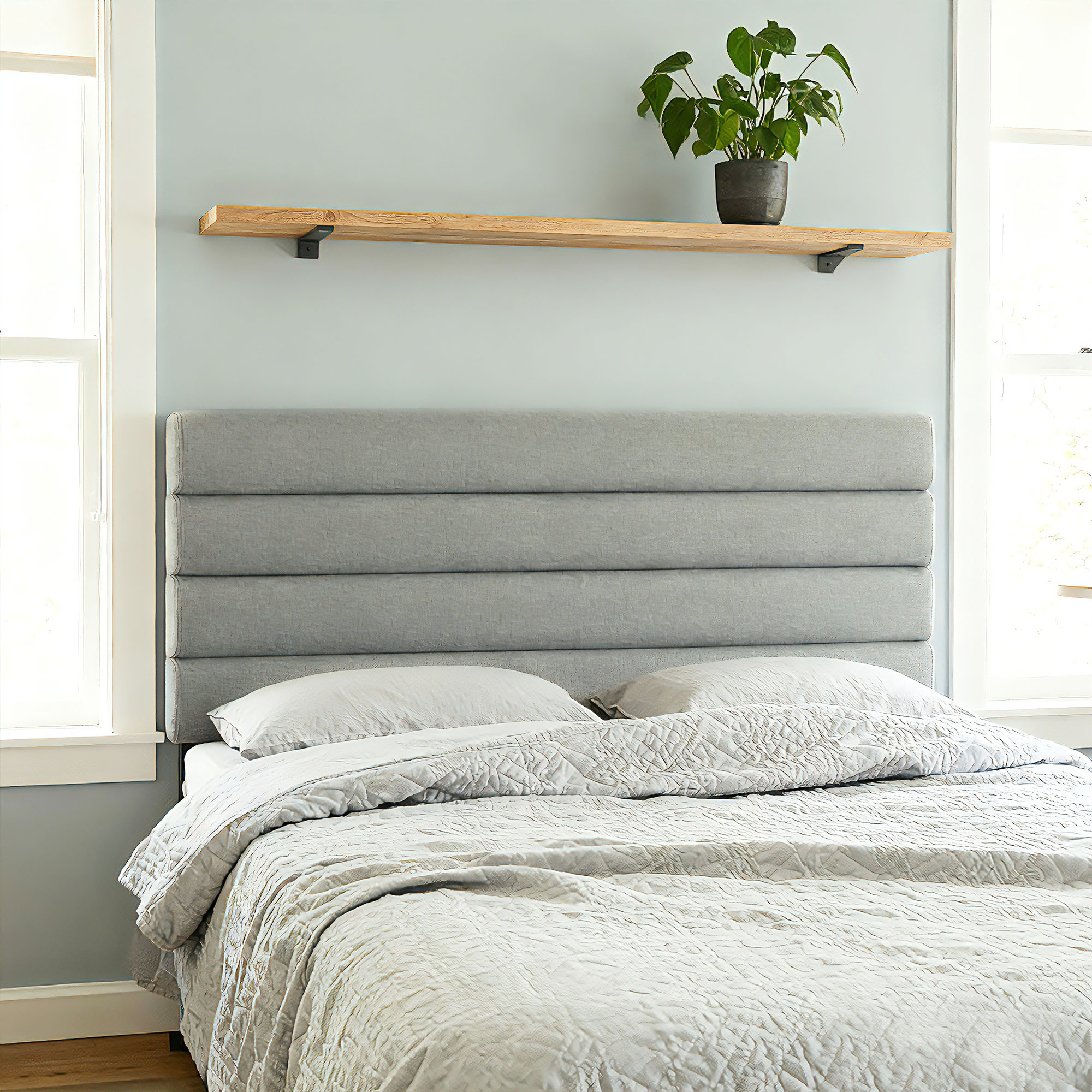 Una cama gris con colcha acolchada y almohadas, junto a una ventana y una estantería con planta.