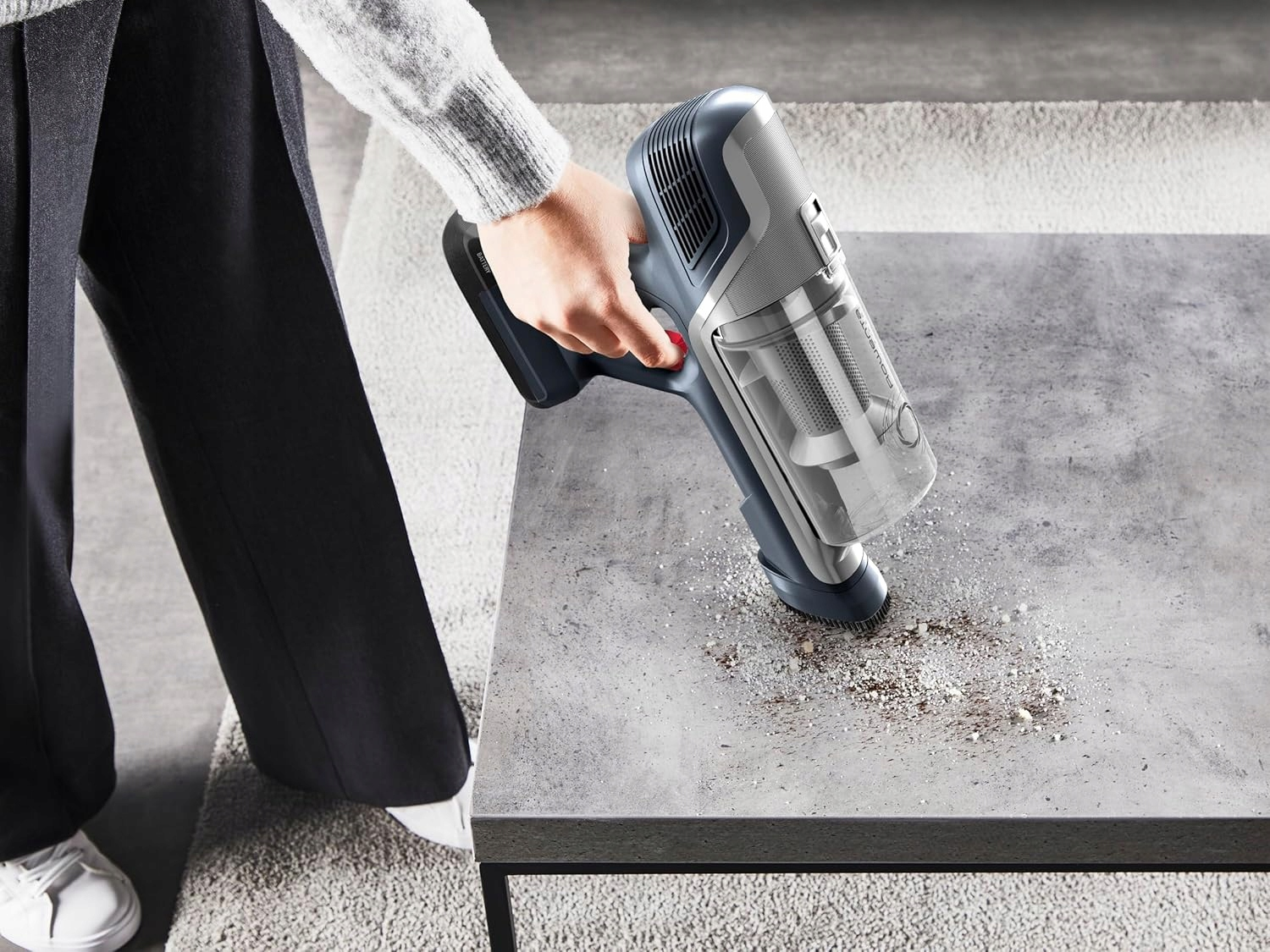 Ręka używa szarego odkurzacza ręcznego do odkurzania brudu z betonowej powierzchni.