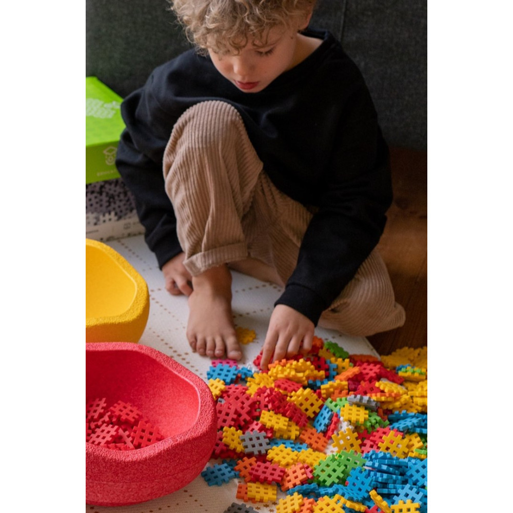 Un niño pequeño juega con bloques de construcción coloridos en el suelo. Dos cuencos, rojo y amarillo, contienen bloques.