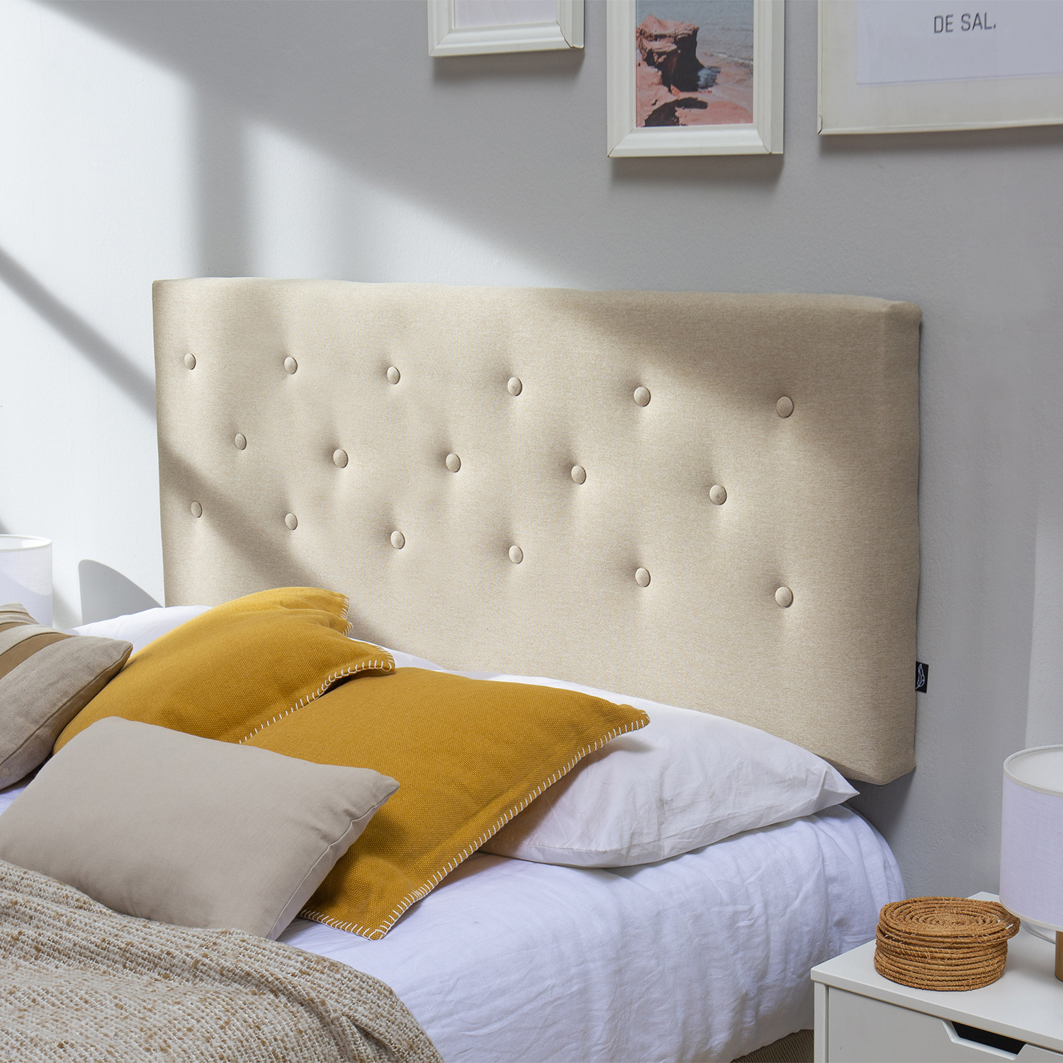 Un cabecero tapizado beige con botones, sobre una cama con cojines amarillos y beige.