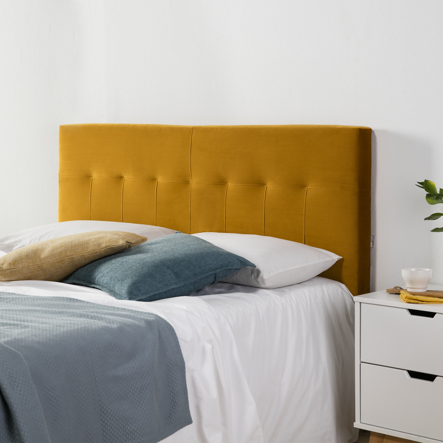 Una cama con cabecero amarillo mostaza, sábanas blancas y cojines azules y amarillos.