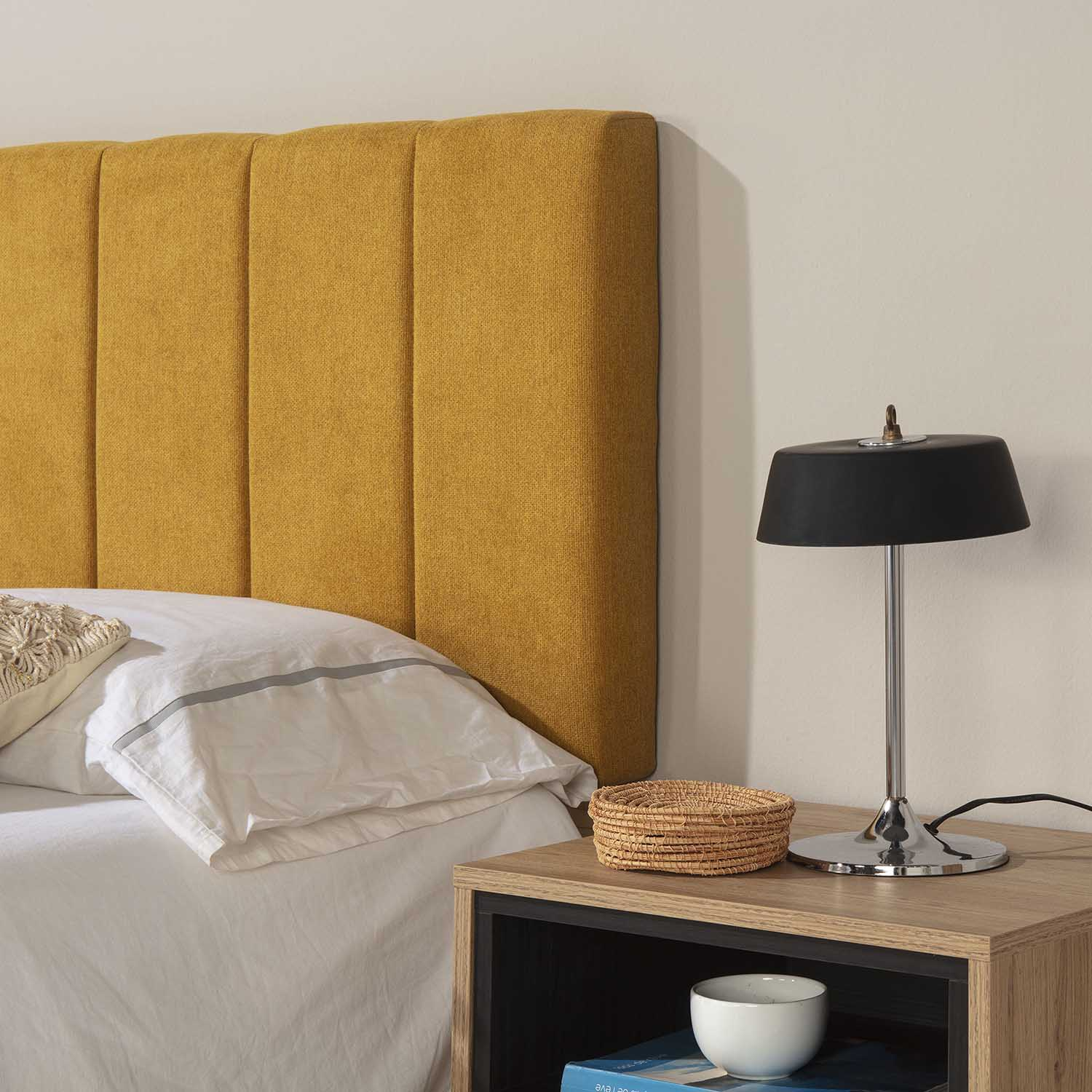 Un cabecero amarillo, mesita de noche con lámpara, cesta y cuenco.