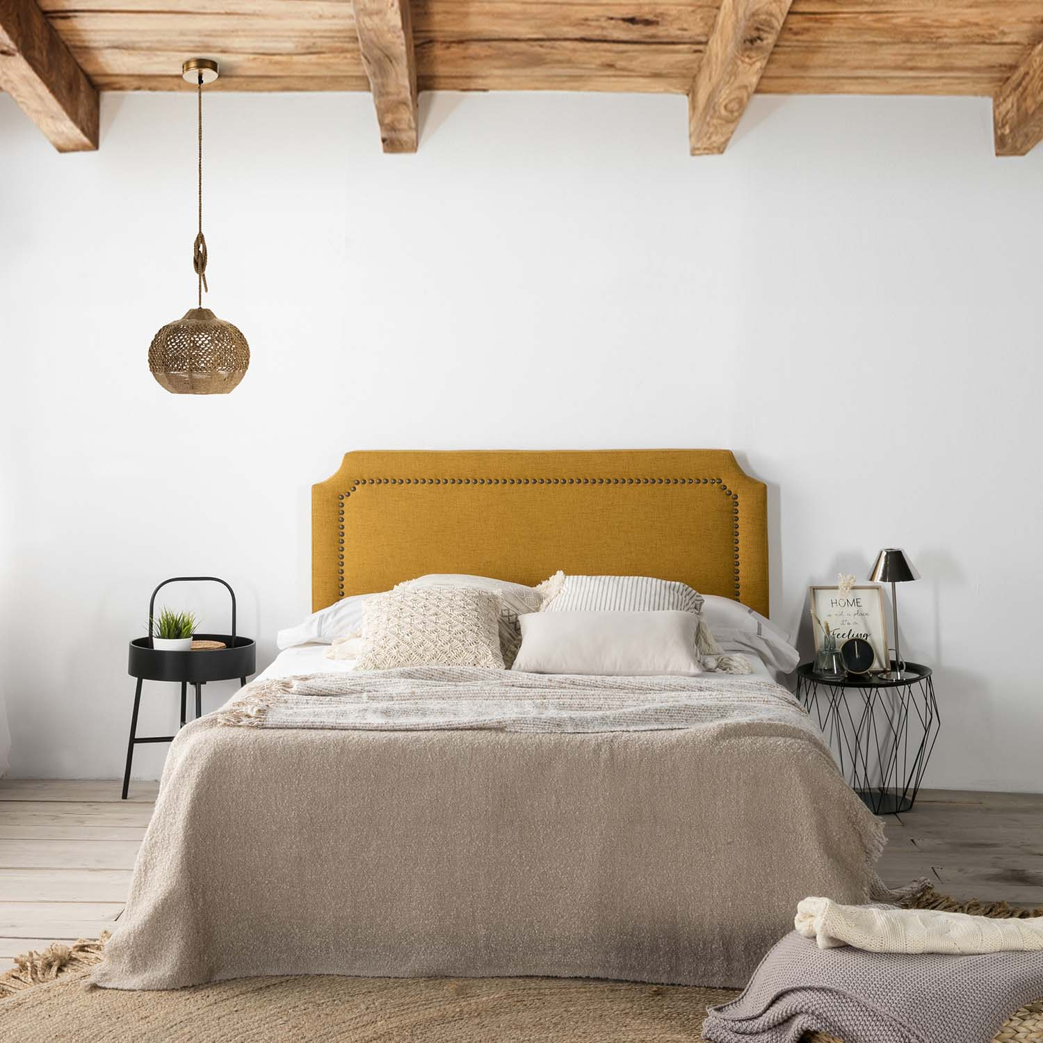 Una cama amarilla con cojines y mantas, mesitas de noche, lámpara y vigas.