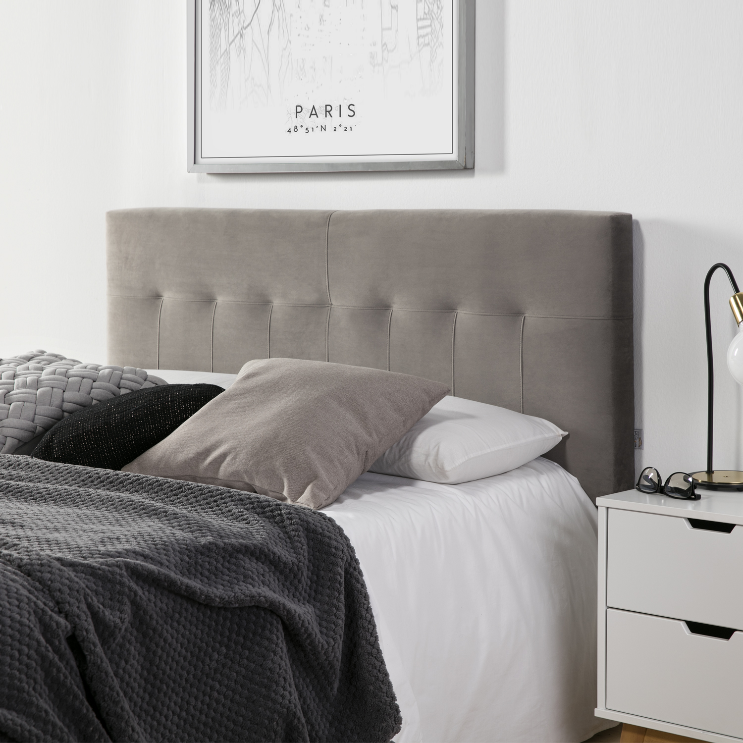 Una cama con cabecero gris, cojines, manta y mesita de noche.